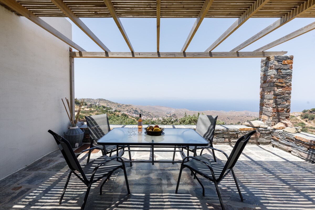 An outdoor dining area is featured under a wooden pergola, overlooking a scenic landscape. A glass table is surrounded by six black chairs. A bowl of fruit is positioned in the center of the table, with views of hills and the Aegean Sea visible in the background.
