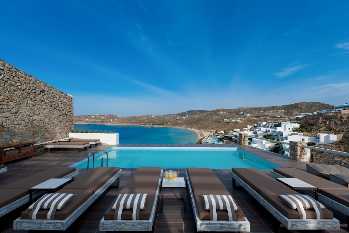 An outdoor swimming pool is visible, surrounded by multiple sunbeds arranged along the deck. A panoramic view of Elia beach and the coastline is featured in the background, under a clear blue sky.