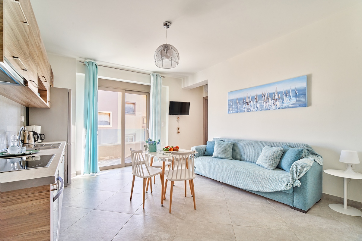 A bright living space features a light blue sofa and a round dining table surrounded by four chairs. Large windows with sheer curtains allow natural light to fill the area. A kitchen is visible in the background, equipped with modern appliances and cabinetry.