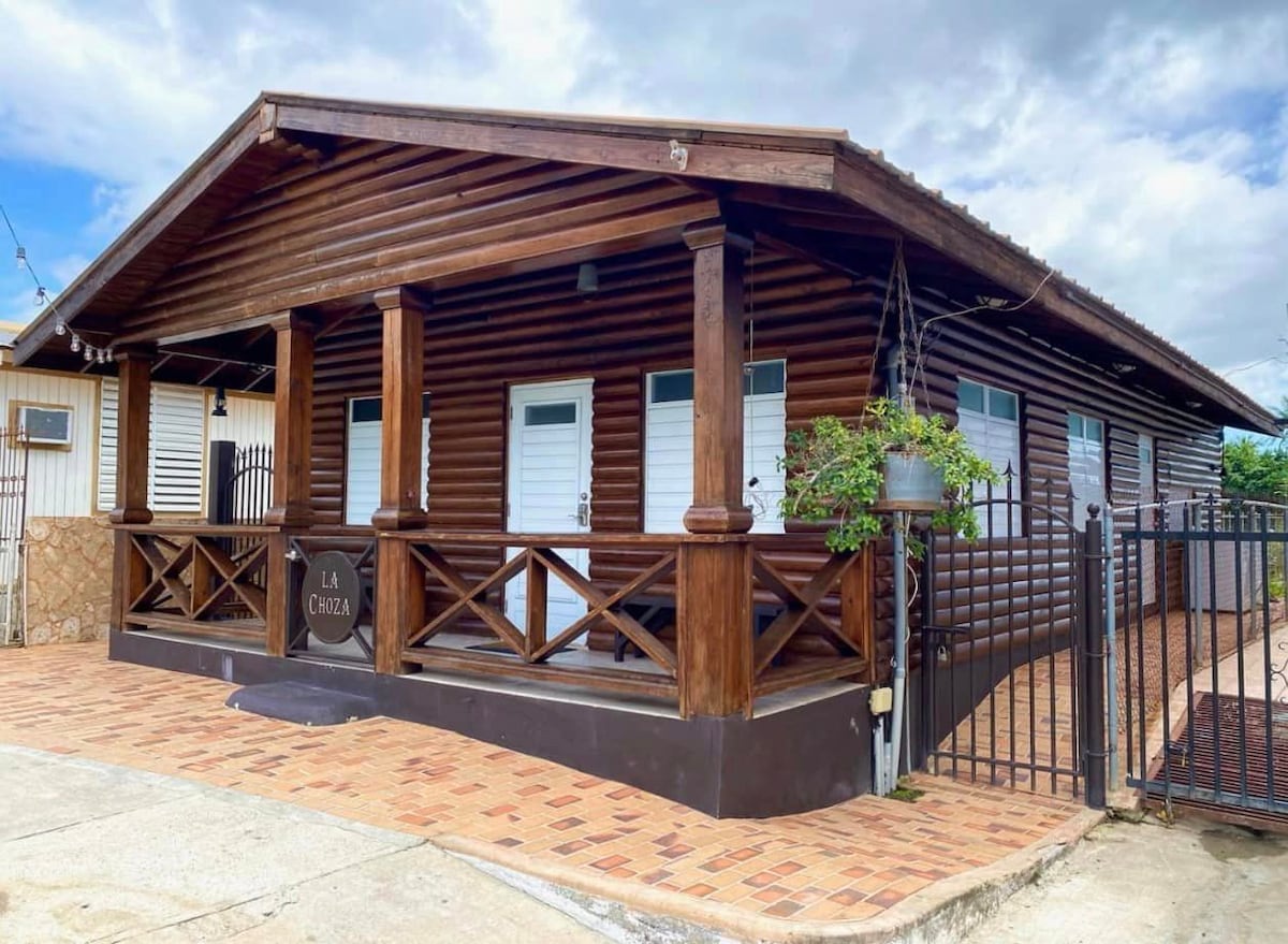 The exterior of La Choza features a rustic wooden structure with a covered porch supported by sturdy posts. Two doors are visible, framed by windows. A paver driveway leads to a gated entrance, and a small plant adds a touch of greenery.