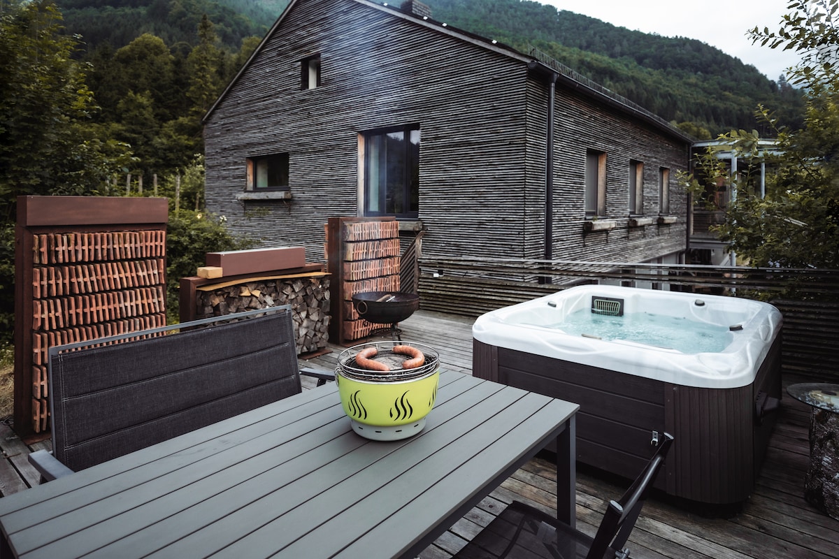 An outdoor area features a private hot tub beside a dining table with four chairs, complemented by a grill and wooden firewood storage. The backdrop includes a rustic building with large windows, set against a green hillside.