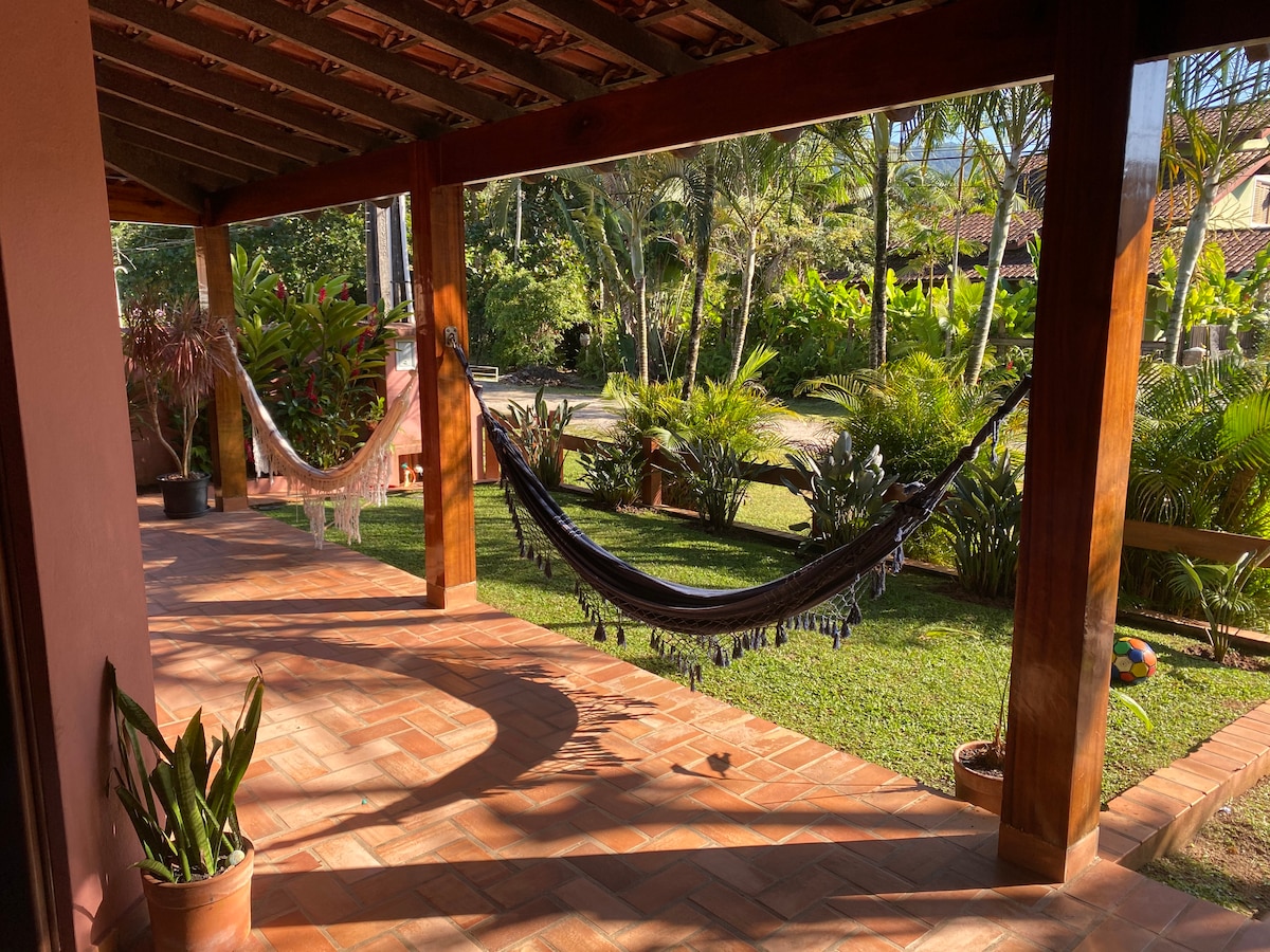 Two hammocks are suspended between wooden posts on a spacious patio, surrounded by lush greenery. The tiled floor is complemented by potted plants, while sunlight casts gentle shadows, creating a relaxed atmosphere. The serene landscape features tropical plants and foliage in the background.