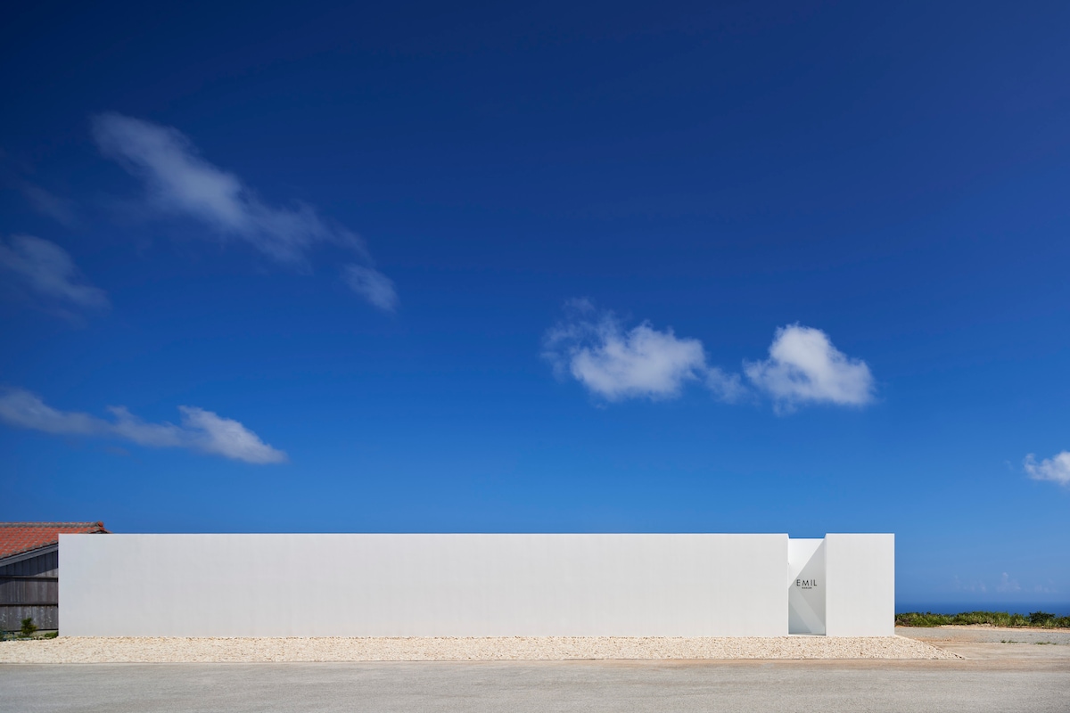 A minimalist exterior of the hotel features a smooth white wall that contrasts with a bright blue sky. The clean lines and simplicity of the design create a serene first impression. A subtle entrance is marked by a small sign on the wall.