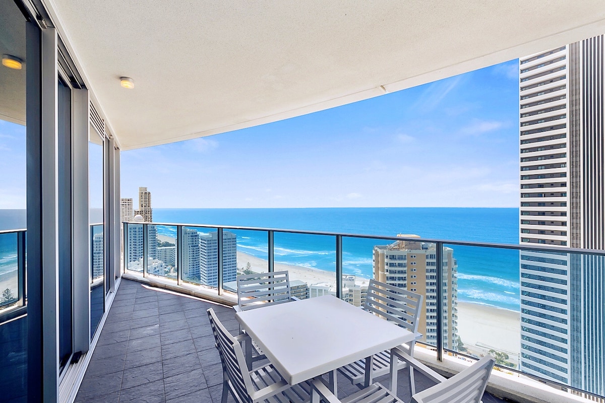 A spacious private balcony offers breathtaking views of the ocean and coastline. Modern outdoor furniture is arranged for relaxation, with clear skies enhancing the beach scenery. Tall buildings frame the view, reflecting the vibrant atmosphere of the surrounding area.