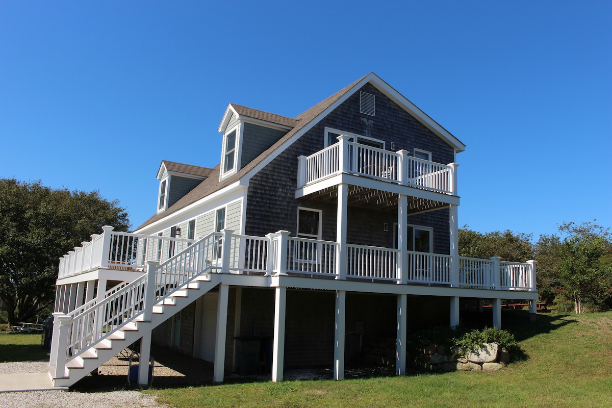 The exterior of the house is showcased with multiple levels and a combination of shingle and clapboard siding. Two spacious decks are featured at the front, complemented by a wide staircase leading up to the entrance. A clear blue sky creates a vibrant backdrop.