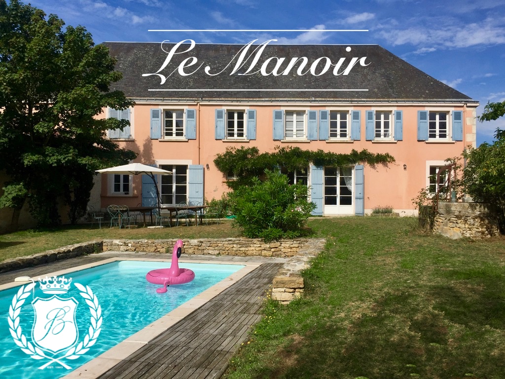 The exterior of Le Manoir is showcased with its pink façade and blue shutters, surrounded by greenery. A private pool with a wooden deck is visible in the foreground, featuring a floating inflatable in the water. Outdoor seating is available under an umbrella to the left.