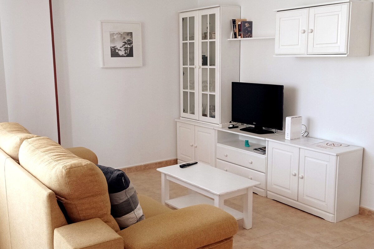 A bright living area is furnished with a beige sofa, a white coffee table, and a TV stand. A television is positioned on the stand, while a glass-front cabinet displays books and decor. Natural light fills the space, highlighting the light-colored walls and tiled flooring.