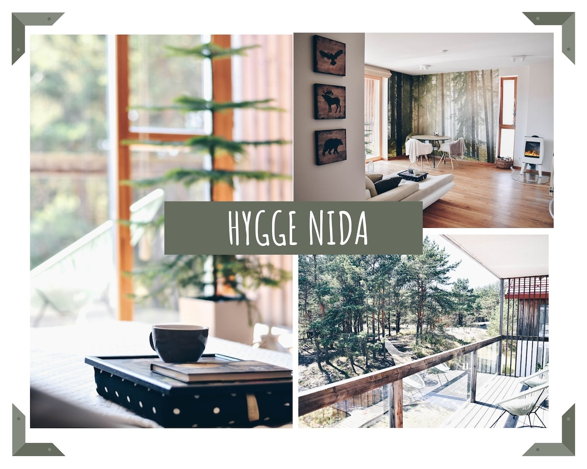 The apartment features wooden floors and large windows that invite natural light. A cozy seating area with a table is visible, alongside decorative wall art. A spacious balcony offers views of surrounding greenery, providing a serene connection to nature.