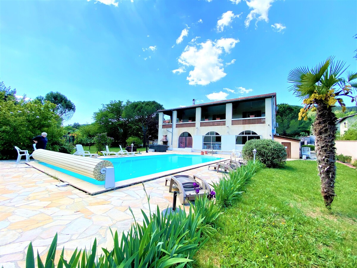 A spacious pool area is bordered by lush greenery and flowering plants, with a blue pool taking center stage. The multi-level villa is visible in the background, featuring large windows and a covered balcony. Several lounge chairs are positioned around the poolside for relaxation.