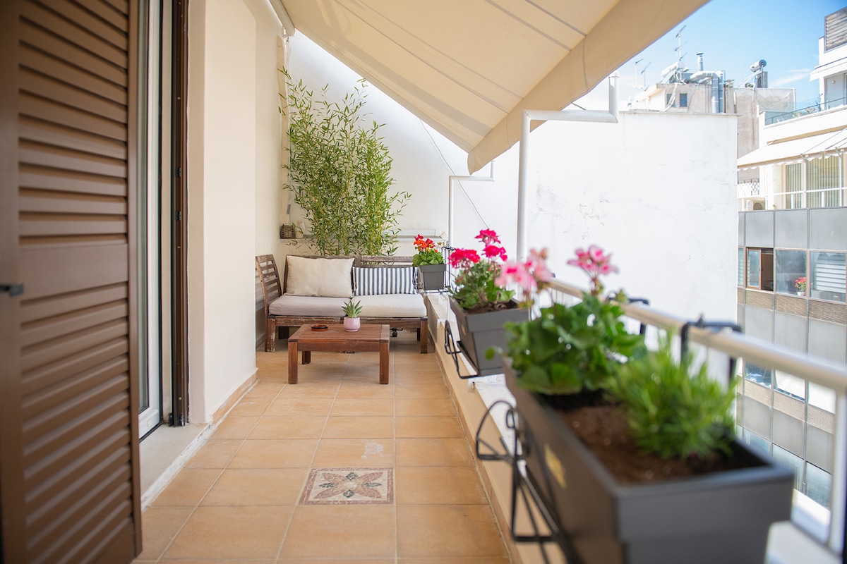 A private balcony is showcased, featuring potted flowers and greenery. A comfortable seating area with a small table is visible, set against a backdrop of a light-colored wall. The balcony is partially shaded by an awning, enhancing a calm outdoor space.