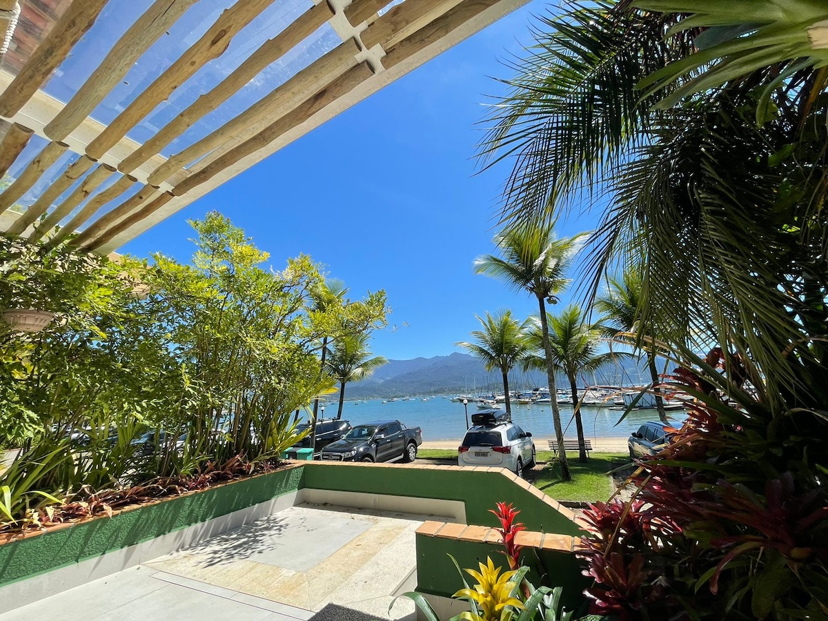 A view from the chalé shows lush greenery framing a clear blue sky. The ocean and a marina are visible in the distance, with a parking area and palm trees enhancing the coastal ambiance.