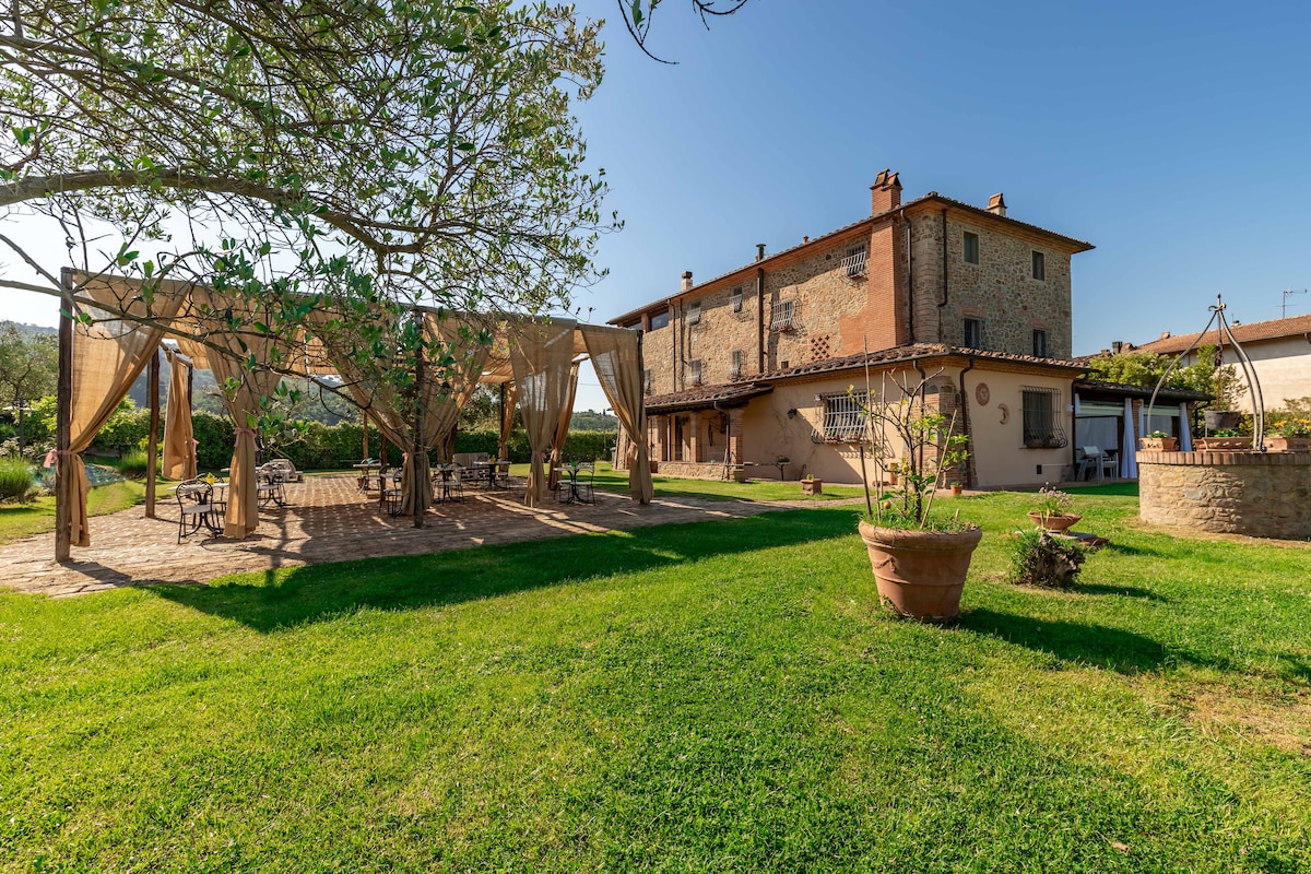 A lush green garden surrounds a charming two-story country house. A shaded seating area with draped fabric is positioned beneath a large tree, offering a place to relax. Potted plants and open lawn spaces enhance the inviting outdoor setting.