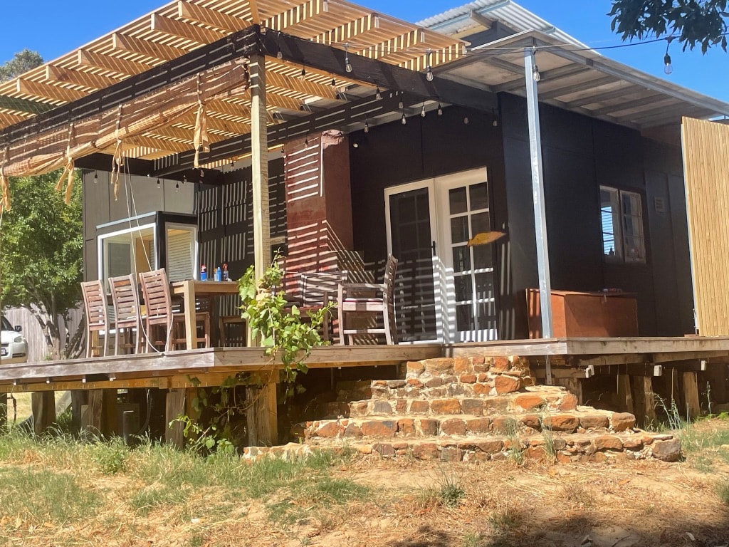 The holiday cabin's exterior features a spacious deck with a dining area and multiple wooden chairs. Sunlight filters through a pergola overhead, providing shade. A stone pathway leads to the entrance, while the structure is surrounded by natural greenery, enhancing the peaceful setting.