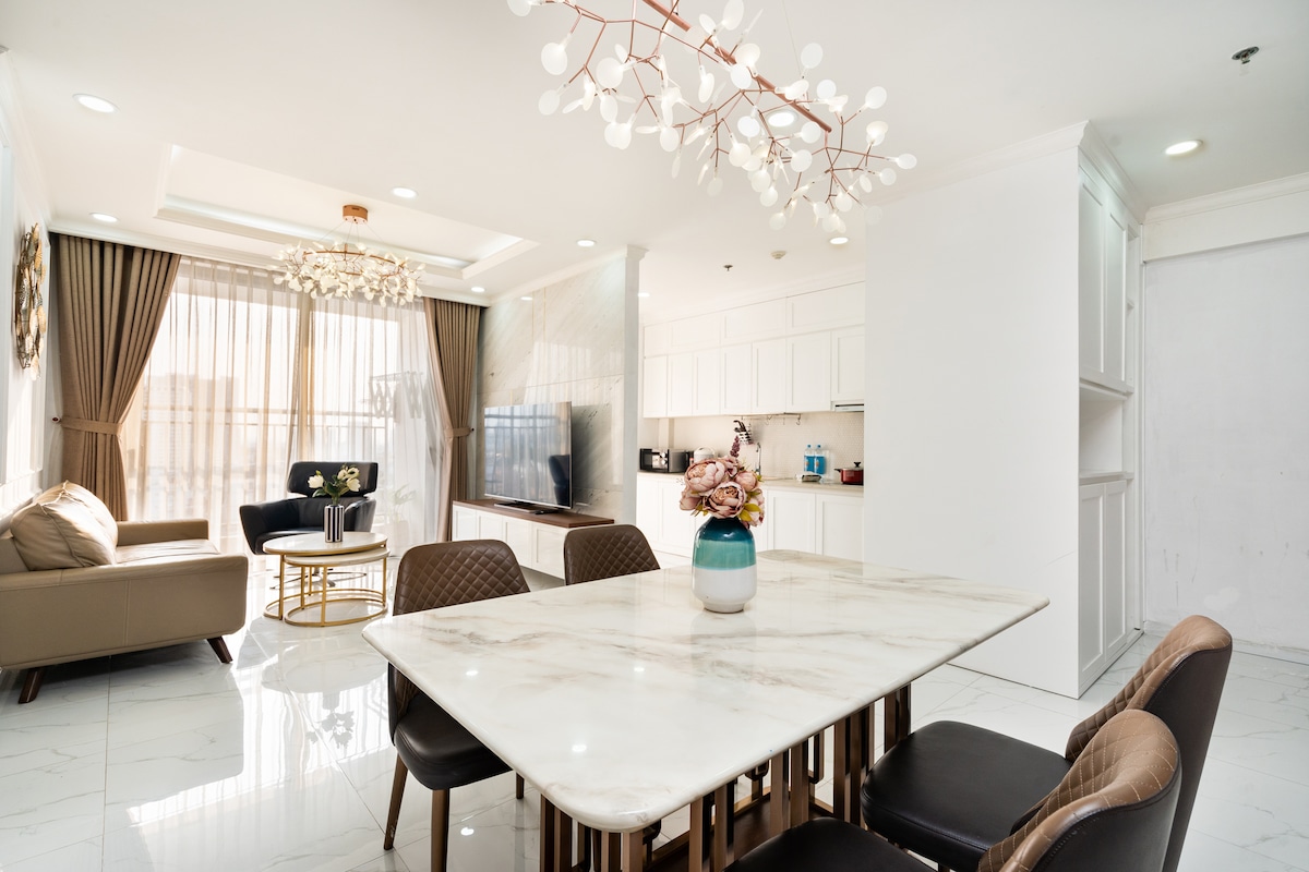 A contemporary living space is shown, featuring a white marble dining table surrounded by upholstered chairs. A cozy sofa and armchair are situated in a well-lit lounge area, complemented by elegant light fixtures and a modern television set, with natural light filtering through large windows.
