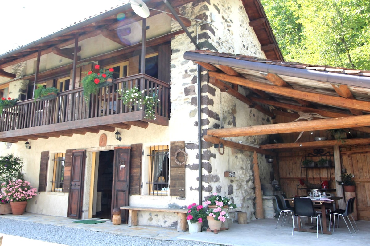The exterior of a stone house showcases a traditional design with wooden accents and flower pots. A covered patio with a dining area is visible, surrounded by greenery. A balcony adorned with flower boxes adds charm to the two-story structure, set against a natural backdrop.