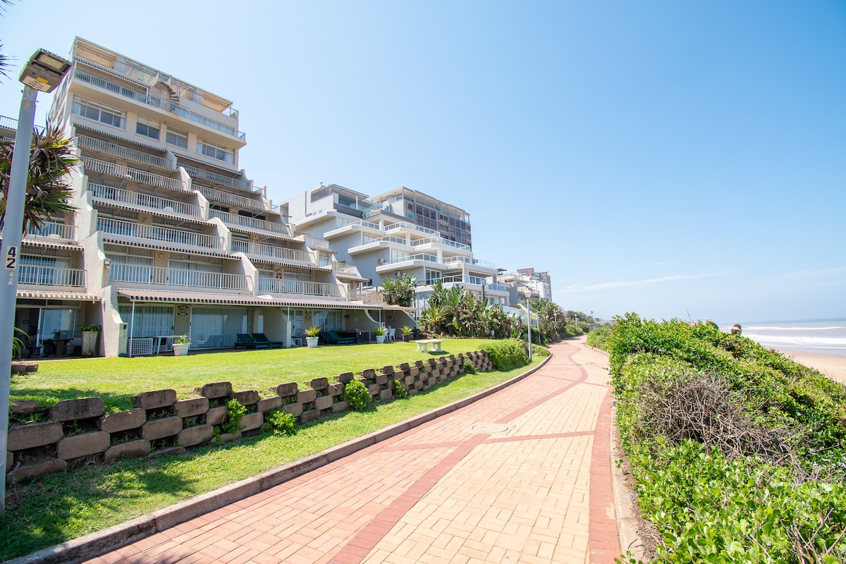 The exterior of a multi-level apartment building is visible, situated along a manicured garden and paved promenade. Lush greenery frames the pathway, which leads towards the beach. The structure showcases multiple balconies and large windows, reflecting a bright, sunny day with clear blue skies.