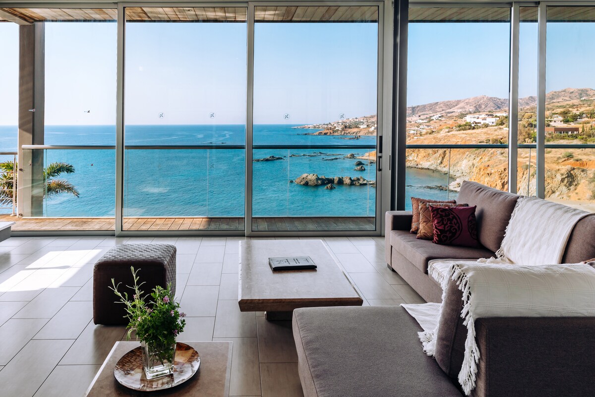 A modern living area features floor-to-ceiling glass walls that provide stunning sea views. A comfortable sectional sofa and a low coffee table create a relaxed setting. Soft natural light fills the space, accentuating the neutral color palette and contemporary design elements.