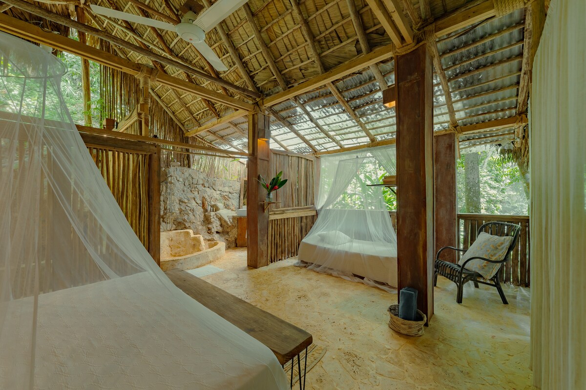 An open, rustic room features spacious beds draped with light, airy canopies. Natural textures in wood and stone create a serene ambiance. Large windows allow soft light to filter through, offering views of lush greenery. A ceiling fan provides a gentle breeze.