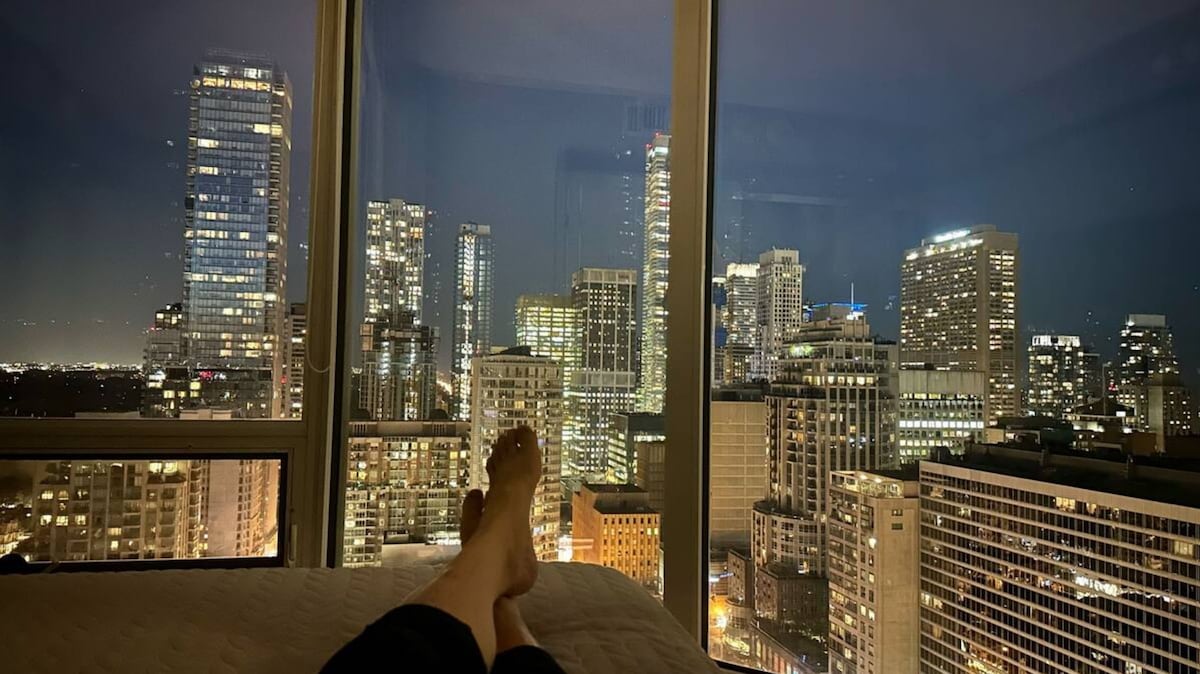 The image showcases a view from the corner suite, featuring a city skyline illuminated at night. Large windows allow for an unobstructed view of the buildings, with twinkling lights creating a vibrant backdrop. A pair of feet rests on the edge of a queen bed, adding a sense of tranquility.