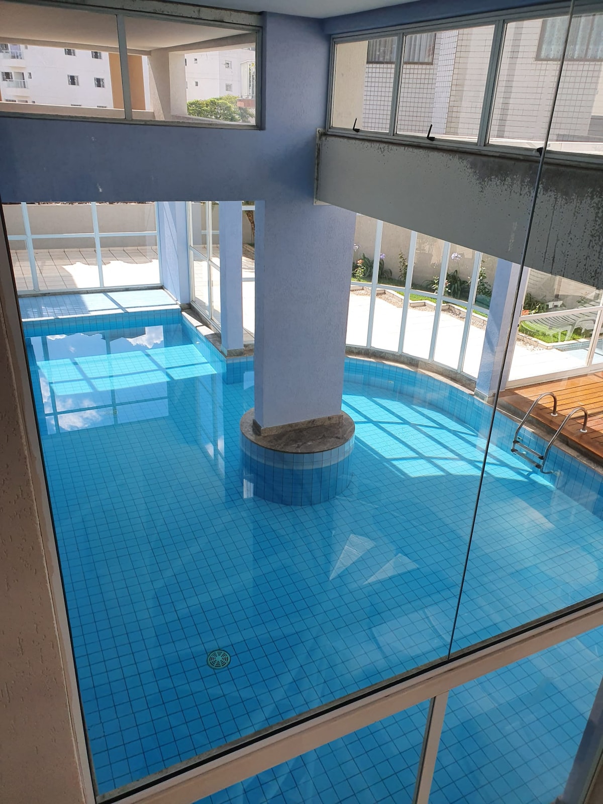 A clear view of the indoor pool is presented, with vibrant blue water reflecting natural light from large windows. Surrounding areas include lounge space, and the pool features a central column. The tranquil atmosphere is enhanced by the airy design and bright surroundings.