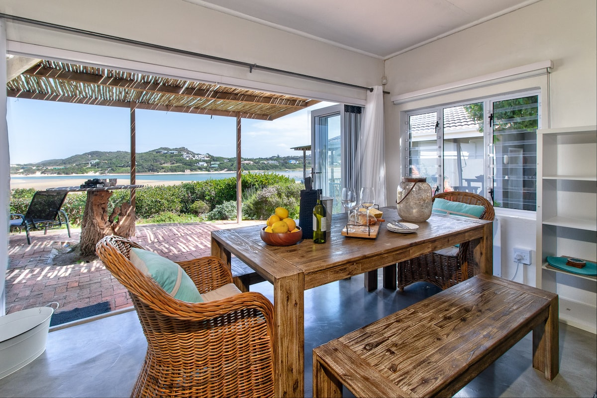 A spacious dining area features a rustic wooden table surrounded by wicker chairs. Sunlight filters through wide-open sliding doors, revealing views of the lagoon and lush greenery. A bowl of fresh fruit, a bottle of wine, and glassware are set on the table, creating an inviting setting.