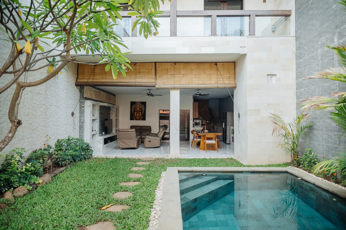 A private swimming pool is nestled beside a well-manicured lawn, surrounded by lush greenery. The villa features an open living area with comfortable seating and a dining table visible through large sliding doors that open onto the patio.