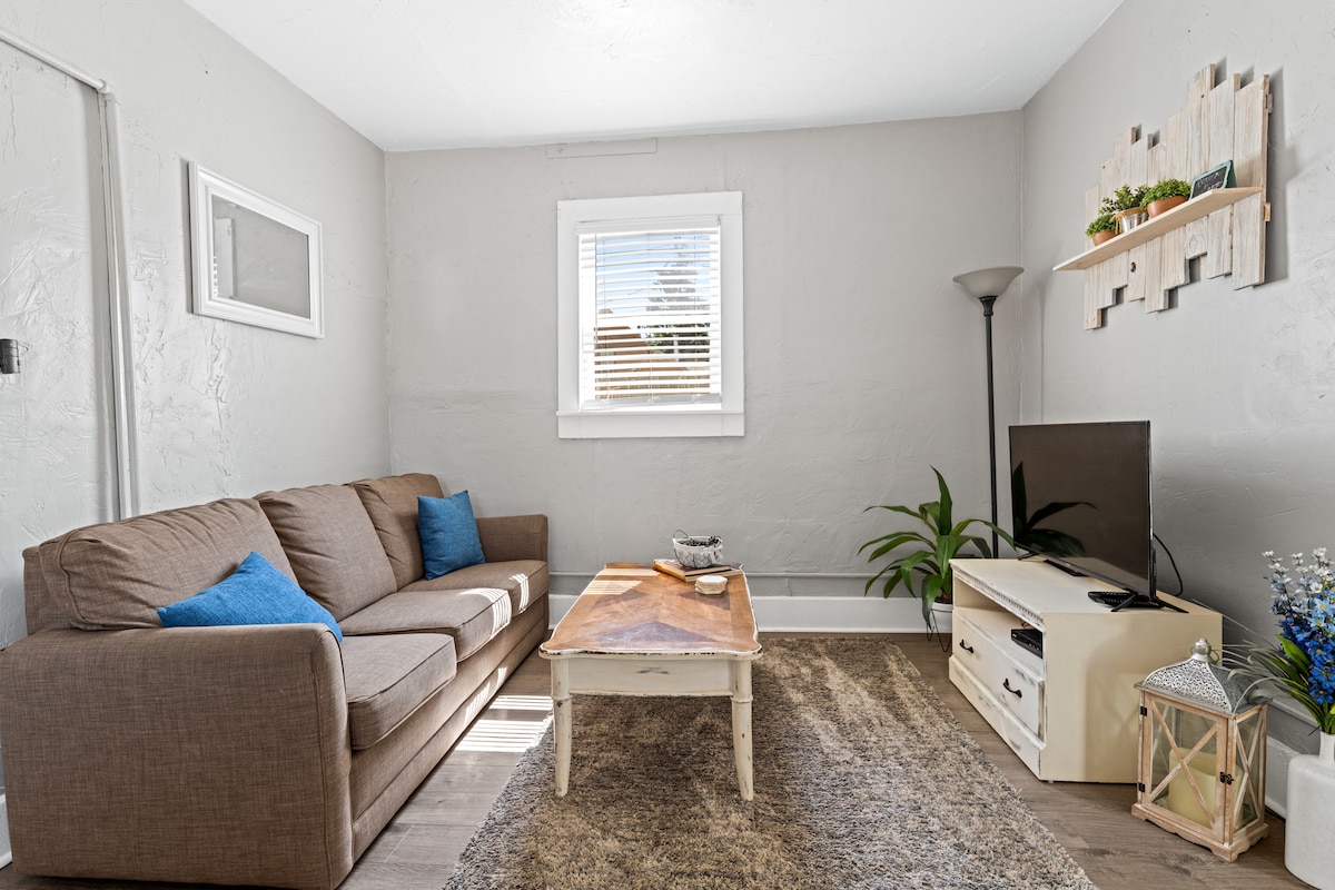 A cozy living area is depicted, featuring a brown sofa adorned with blue accent pillows. A wooden coffee table occupies the center, and a flat-screen TV is positioned on a light-colored console. Natural light flows in through a window, enhancing the welcoming ambiance.
