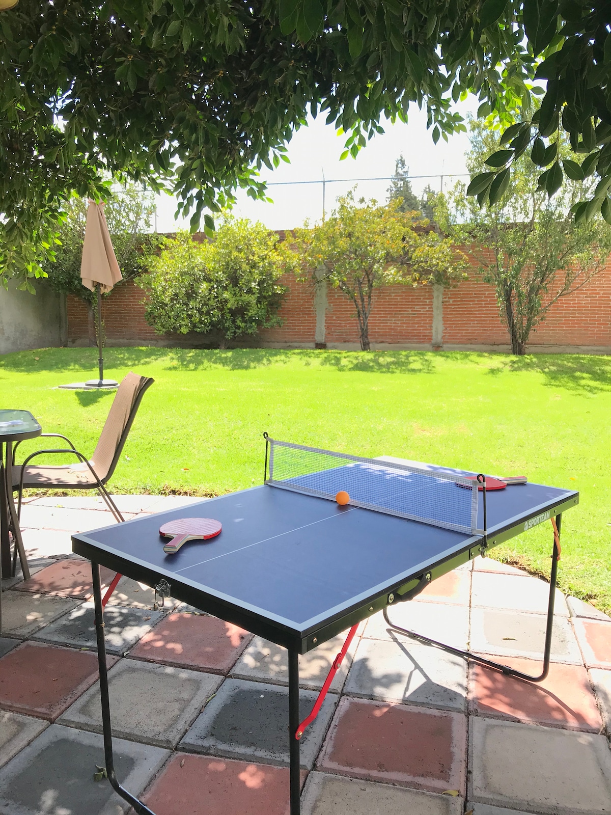 An outdoor table tennis setup is positioned on a stone-paved patio, surrounded by a well-maintained lawn. Sunlight filters through leafy trees in the background. The table holds a net, a ping pong ball, and paddles, inviting guests to enjoy recreational activities in a tranquil setting.