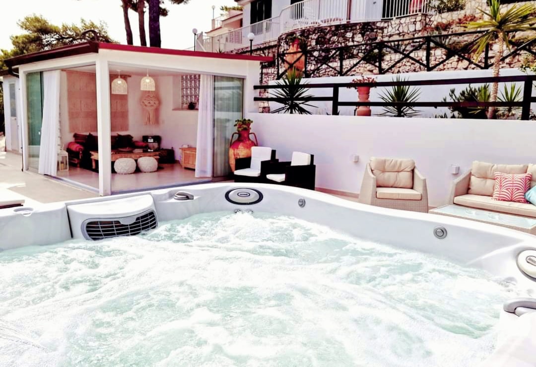 A private jacuzzi spa is shown with bubbling water, surrounded by a relaxing outdoor seating area featuring cushioned chairs and a small table. In the background, a modern glass-enclosed space is visible, enhancing the overall ambiance with plants and natural light.