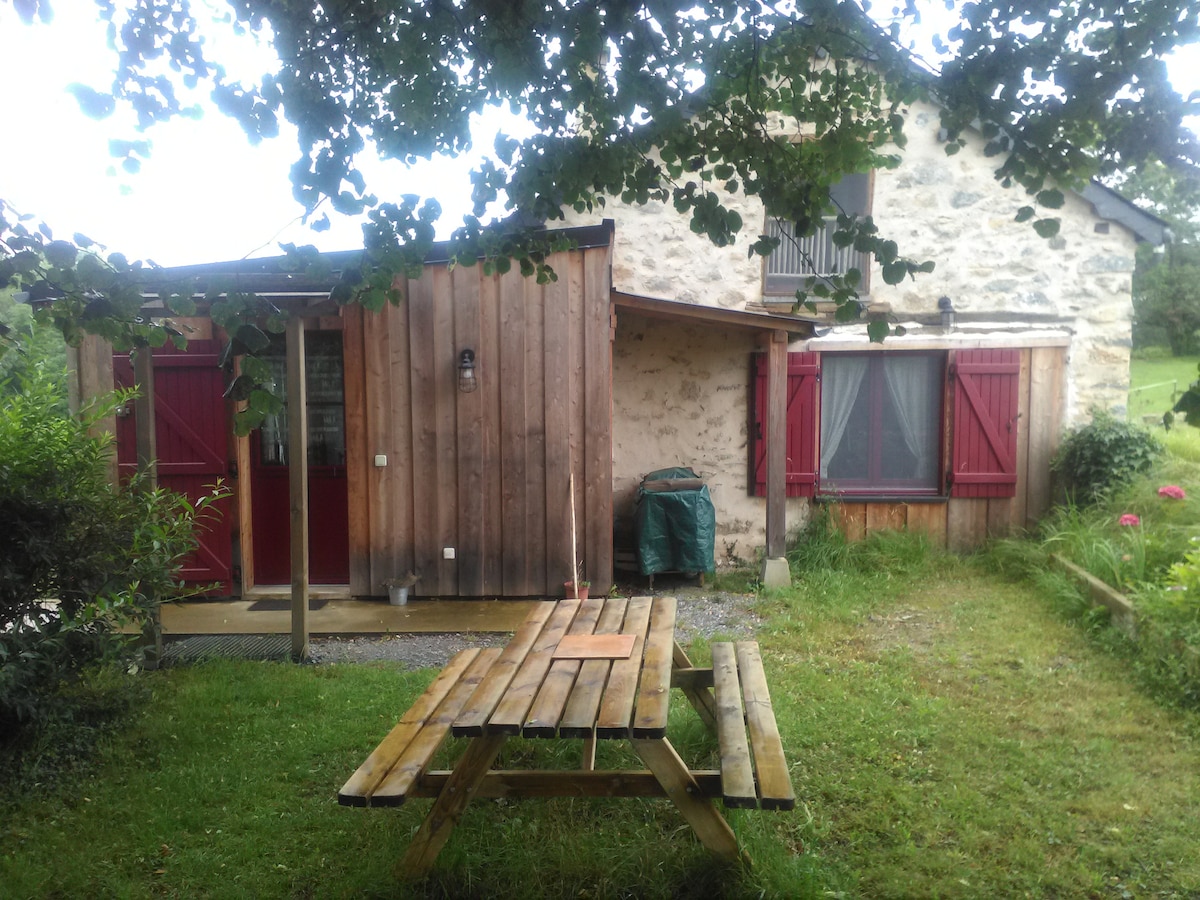 The exterior of the gîte is shown, featuring a charming stone facade accented by wooden panels. A picnic table is situated on the grassy area in front, surrounded by greenery. Red shutters frame the windows, and there is a small green storage unit nearby.