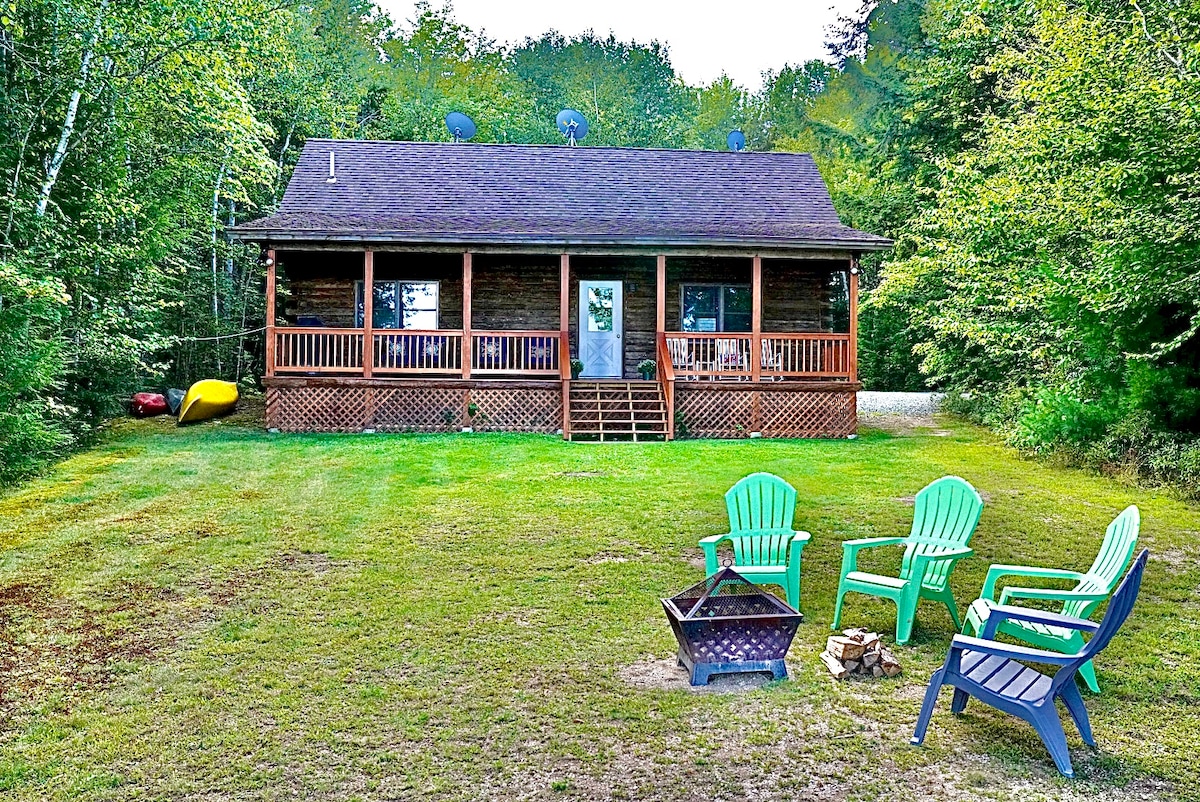 The cabin is surrounded by lush greenery, featuring a welcoming front porch with steps leading up to it. In the foreground, colorful chairs are arranged around a fire pit, complemented by a canoe resting nearby. Natural light illuminates the scene, enhancing the tranquil setting.
