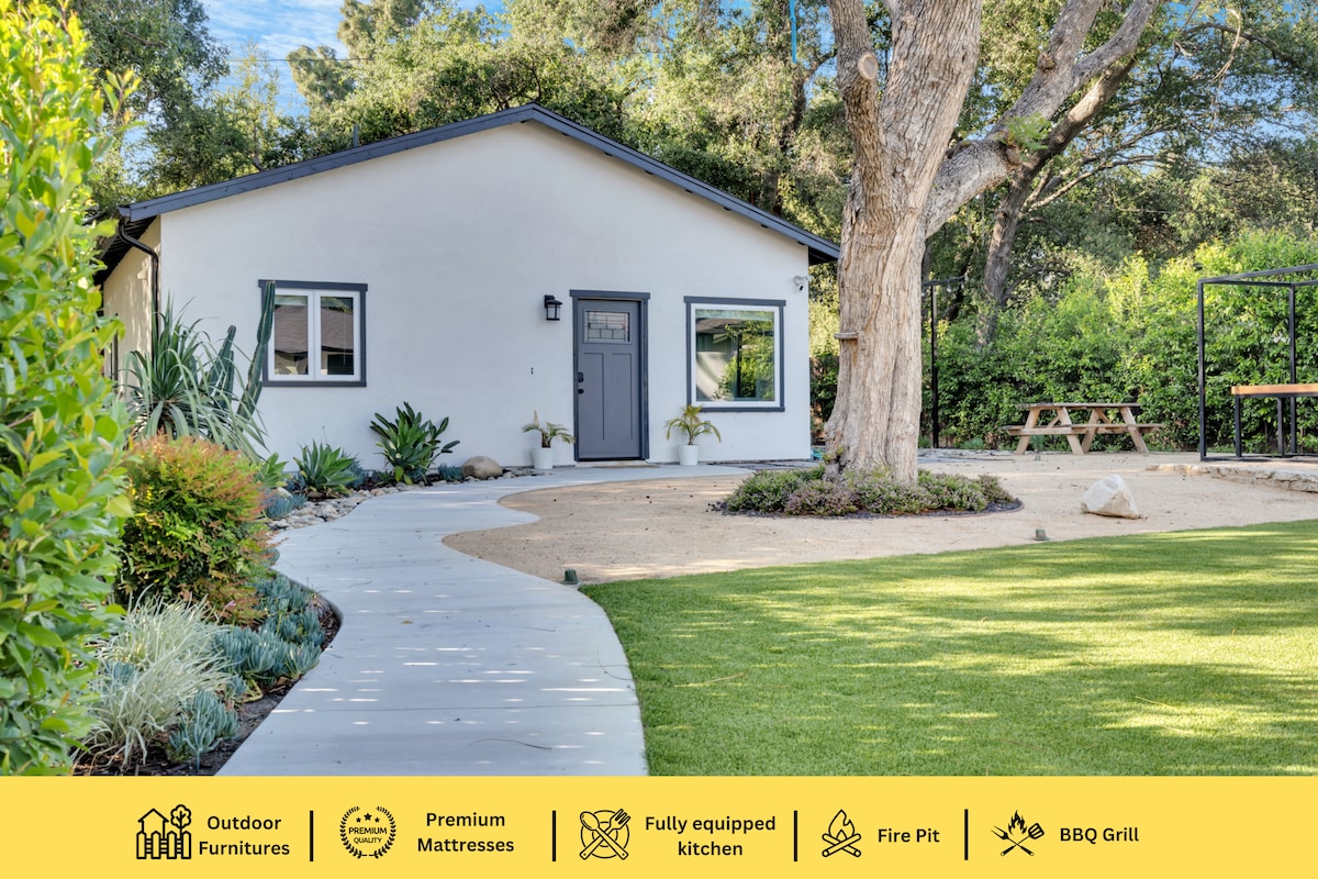 The exterior of the guest house is depicted, featuring a simple white façade and a welcoming front door. The garden is landscaped with greenery, surrounding a path that leads to a yard with outdoor furniture and a fire pit, creating an inviting outdoor space.