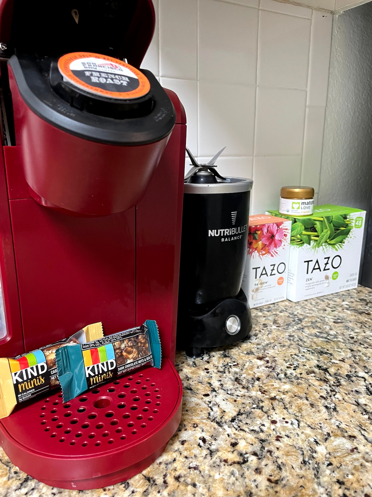 A vibrant coffee maker is set against a light-colored backsplash, with a selection of tea boxes displayed nearby. Snack bars are placed on the counter, showcasing a thoughtful arrangement of refreshments available for guests.