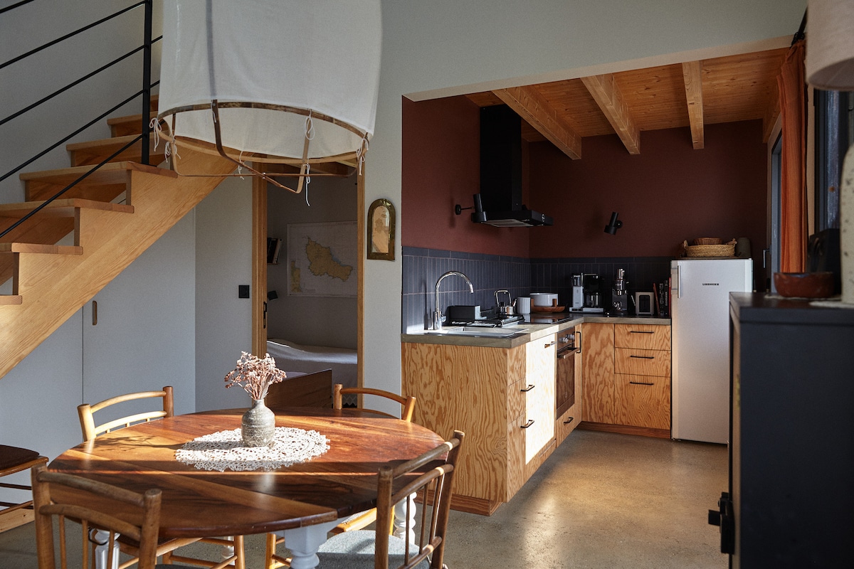 An open-concept living area showcases a wooden dining table surrounded by four chairs, with a decorative centerpiece. The kitchen features modern appliances, wood cabinetry, and a dark tiled backsplash. Natural light floods the space, highlighting the wooden beams of the ceiling.