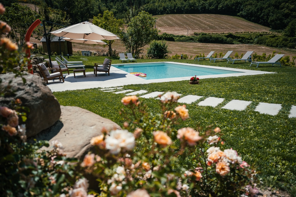 A refreshing outdoor space includes a swimming pool surrounded by lounge chairs and sun umbrellas. Neatly arranged stone pathways lead through a well-maintained lawn, adorned with blooming flowers. Lush greenery and gently rolling hills are visible in the background.