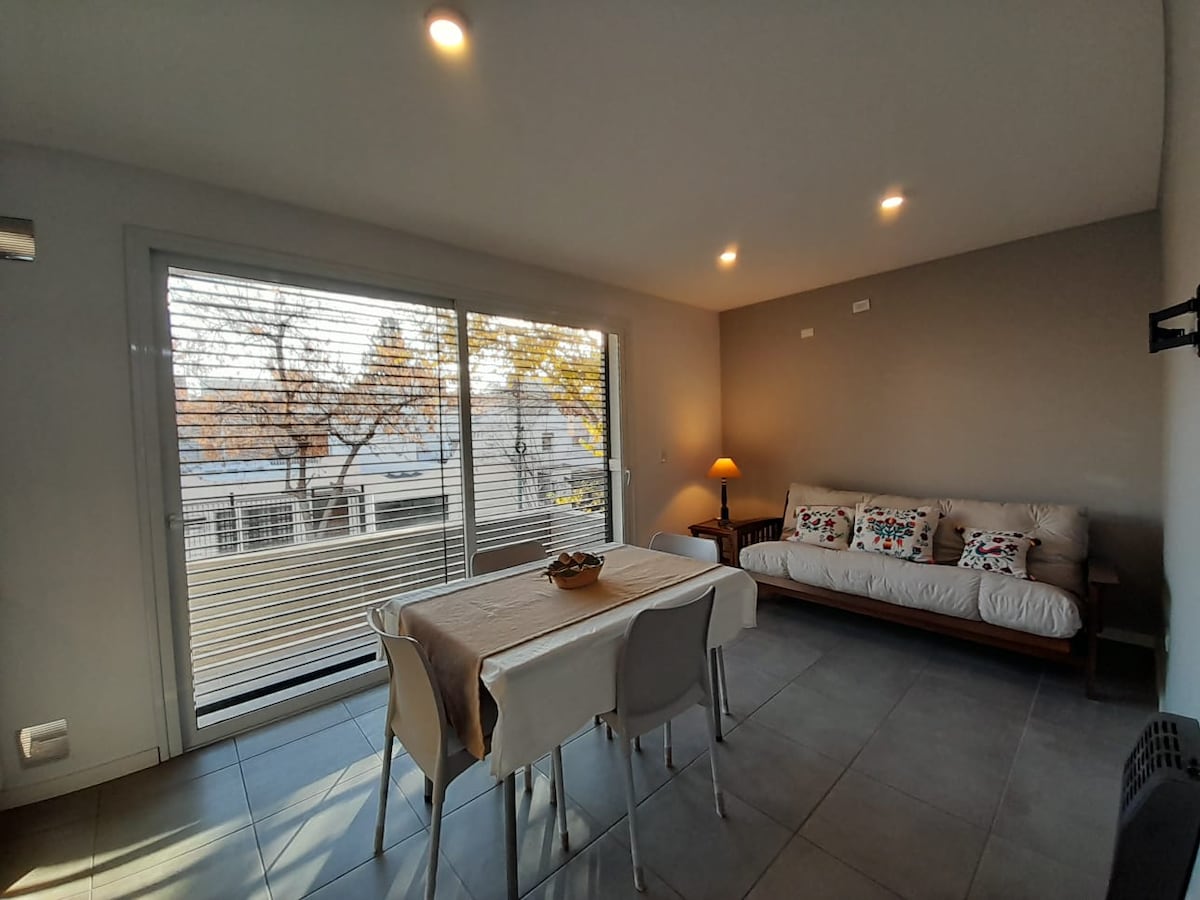 A bright living area features a comfortable sofa adorned with colorful decorative pillows. A simple dining table with four chairs sits nearby. Large windows allow natural light to fill the space, showcasing views of trees outside.