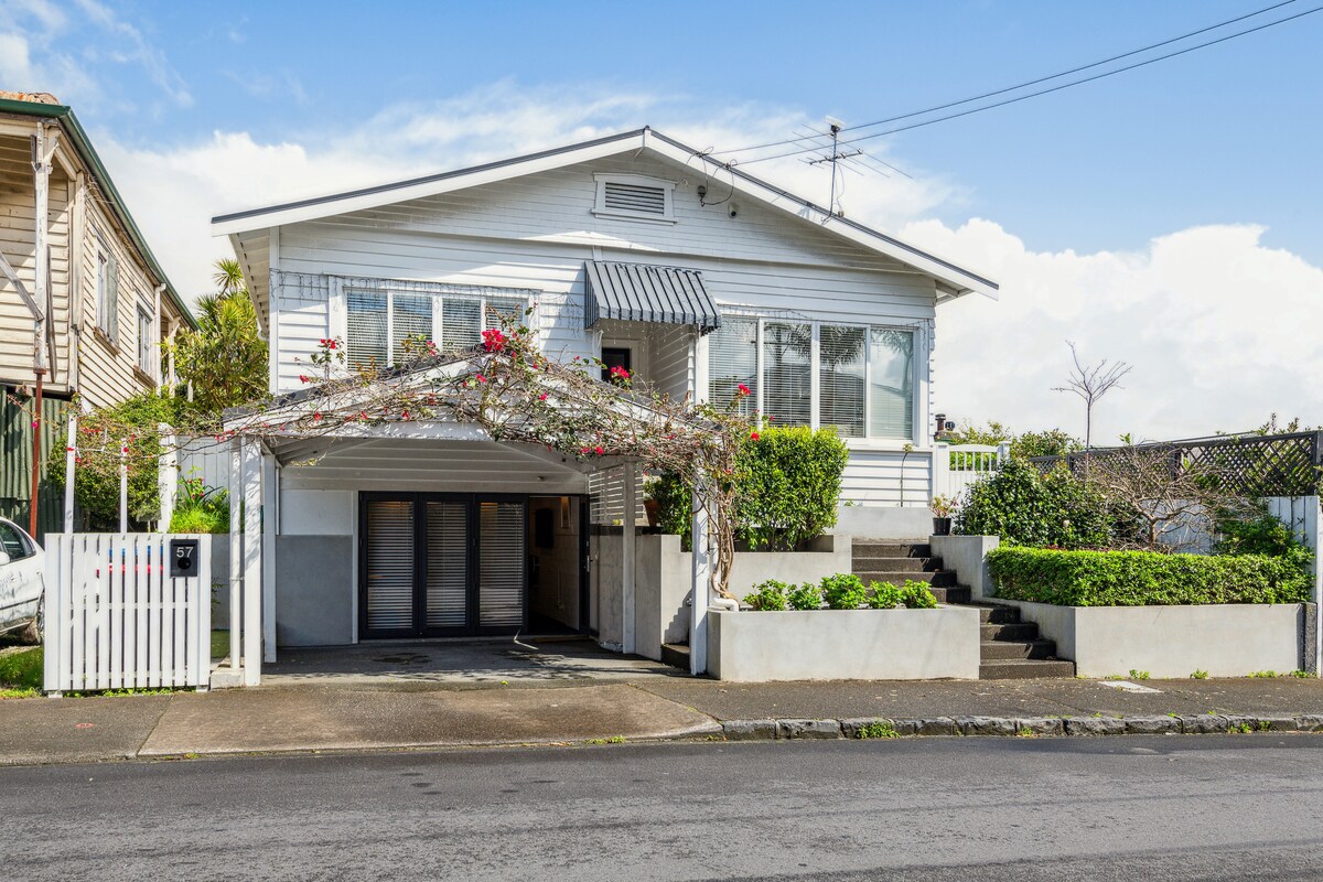 A charming two-story house is shown, featuring a carport adorned with flowering vines. The main entrance is highlighted by a canopy and surrounded by well-maintained greenery. The neighborhood setting indicates accessibility to nearby streets and amenities.