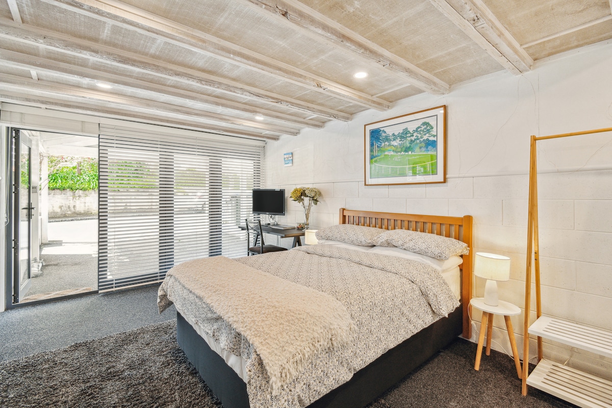 A spacious room features a comfortable double bed with a textured blanket. Natural light filters through large sliding doors that lead to the outside. An office desk with a chair, a small Smart TV, and a stylish lamp are visible, while light wooden beams accent the ceiling.