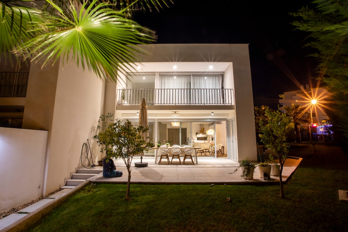 An outdoor terrace extends from the back of the villa, featuring a dining table surrounded by chairs, ideal for gatherings. Soft lighting enhances the ambiance, while tropical plants add a natural touch. The space looks out onto a well-maintained lawn, conveying a sense of serenity.