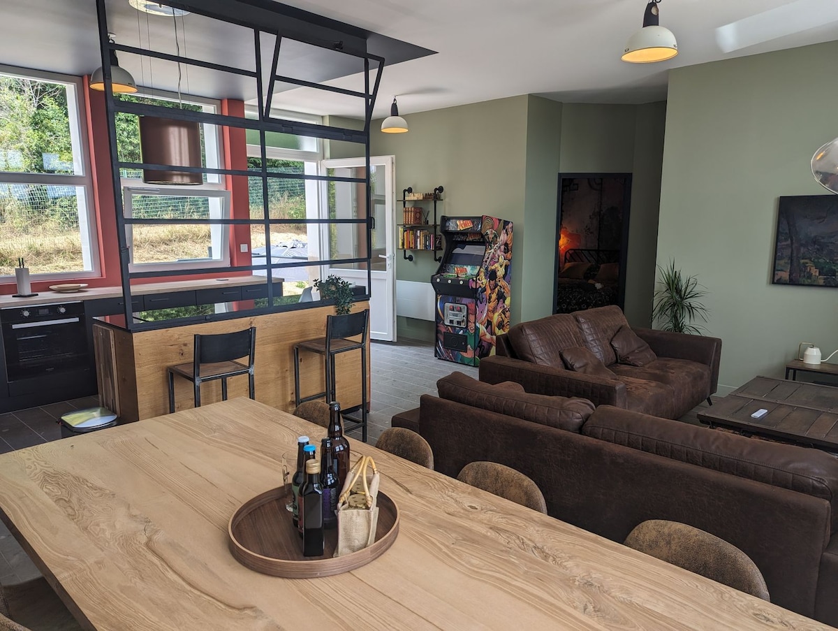An open-plan living area features a spacious dining table with a circular tray containing drinks. A comfortable seating arrangement is highlighted by brown sofas and a large window providing natural light. Nearby, a kitchen area and gaming machine add to the leisure aspects of the space.