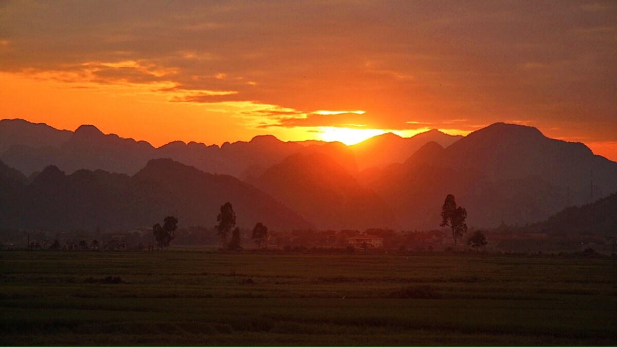 A panoramic view of mountains during sunset, with warm hues of orange and yellow illuminating the landscape. Silhouettes of trees and hills are visible against the vibrant sky, creating a tranquil setting.