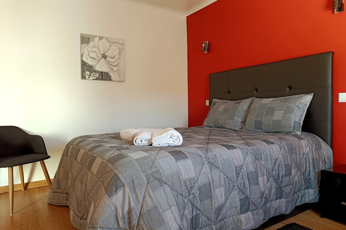 A spacious bed dressed in a textured blanket is positioned against a vibrant red wall. A monochromatic artwork hangs above the bed, and soft, natural light enters from an unseen source. A minimalist chair complements the room's decor.