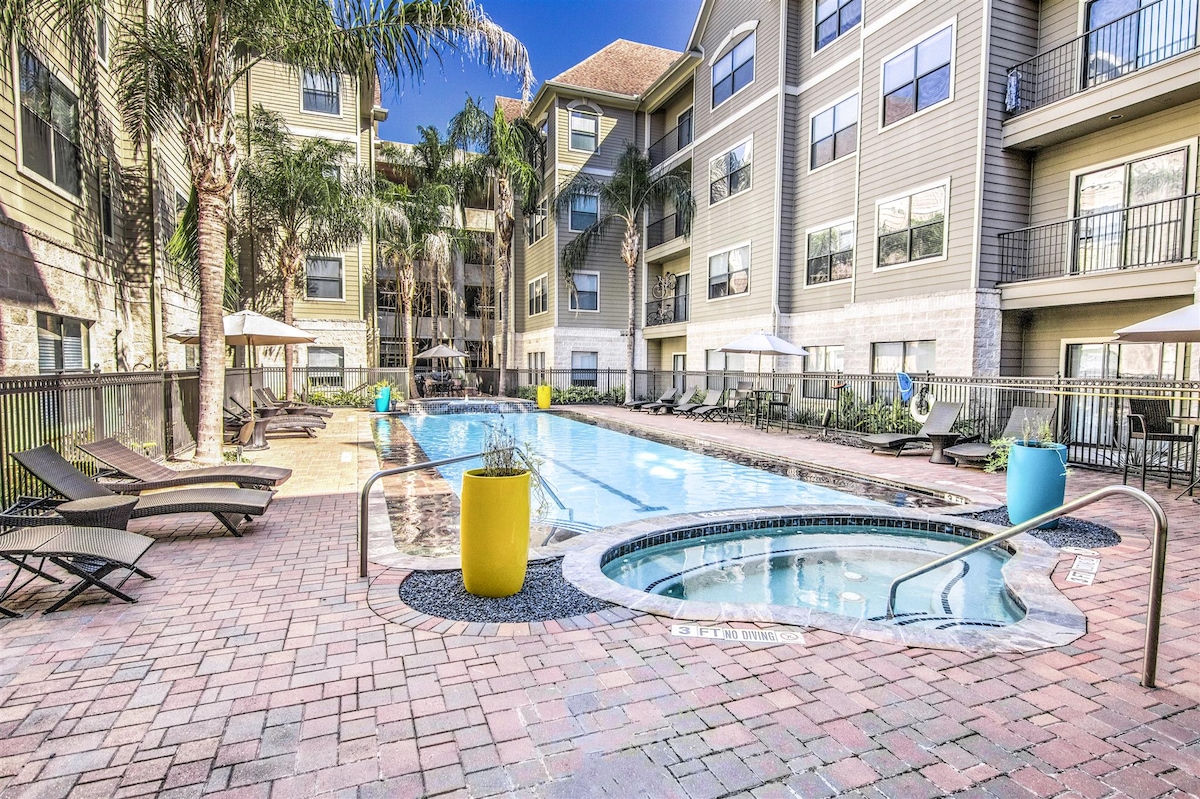 A spacious outdoor area features a resort-style lap pool surrounded by lounge chairs and palm trees. A hot tub is included, with decorative planters providing pops of color. The pool area is bordered by buildings featuring numerous large windows, allowing natural light to flow in.