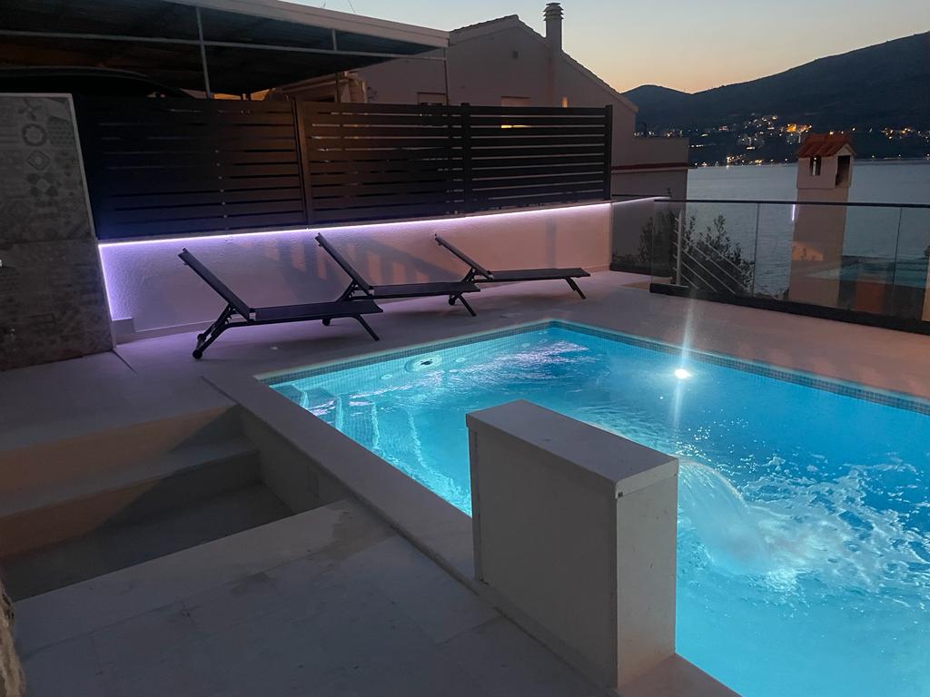 A modern swimming pool is lit with gentle illumination, reflecting the evening sky. Two loungers are positioned nearby, creating a relaxing space for guests. The tranquil water features a built-in massage area, and a glimpse of the surrounding landscape is visible in the background.