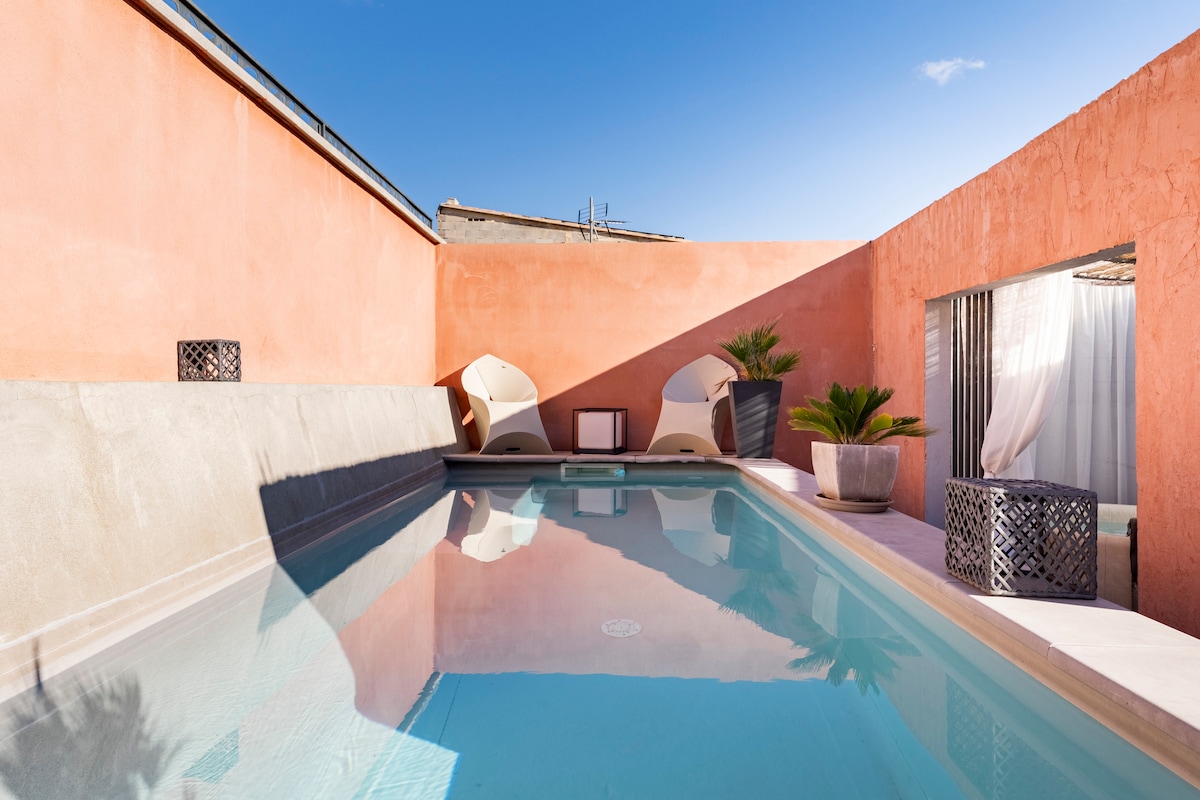 A serene private pool is surrounded by smooth, textured walls in soft pink tones. Comfortable loungers are positioned nearby, offering relaxation opportunities. Sunlight reflects off the clear water, enhancing the tranquil outdoor space, ideal for unwinding.