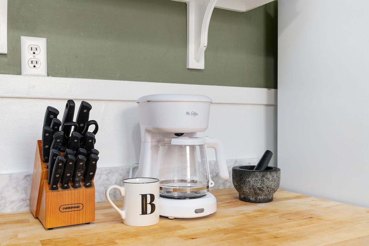 A well-organized kitchen counter displays a coffee maker with a glass carafe, a mug featuring a bold letter, and a mortar and pestle. A wooden knife block with multiple knives is positioned nearby. The backdrop includes textured green walls, contributing to an inviting culinary space.