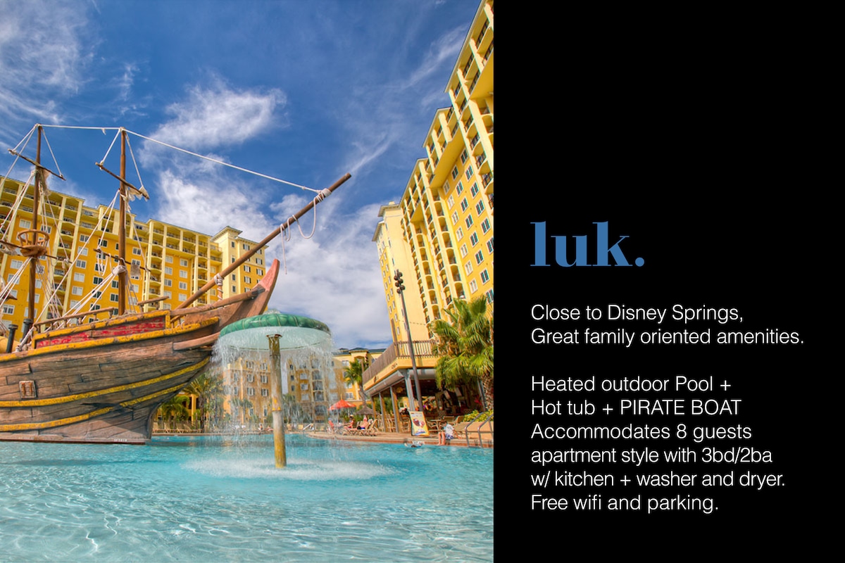 An outdoor area features a heated pool with a pirate boat structure, complemented by a hot tub and cascading water features. Surrounding buildings exhibit a family-friendly resort design, with ample seating available near the pool, and vibrant colors adding a playful touch.