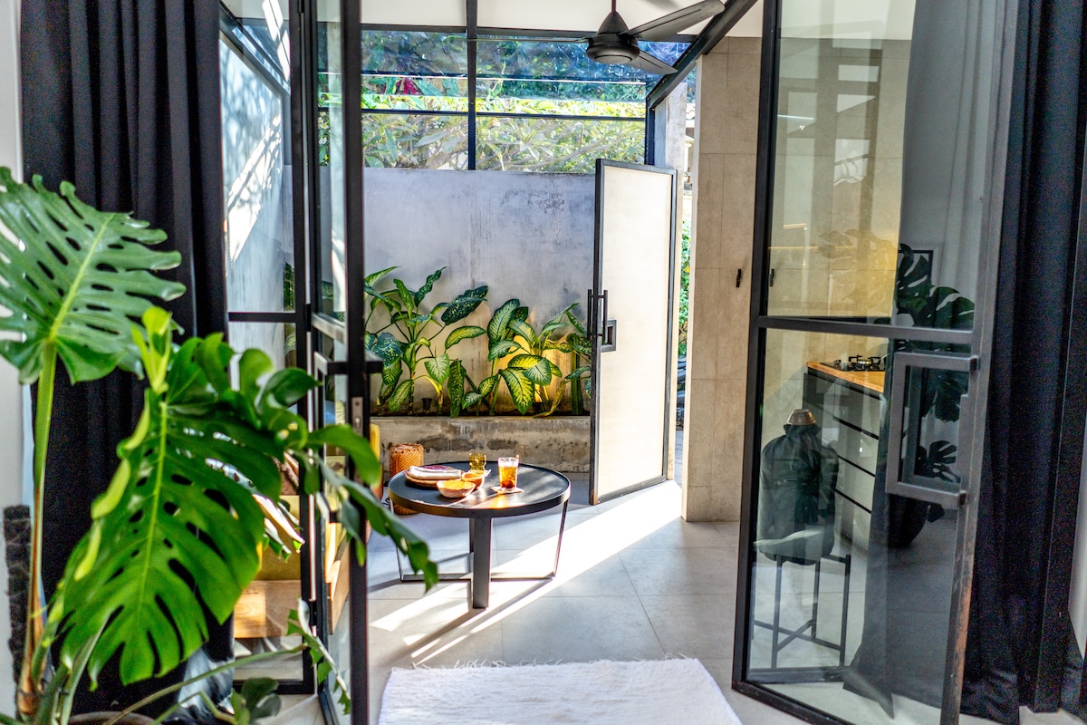 Natural light floods the open space through large glass doors, showcasing a modern kitchen area and a small table set for refreshments. Lush green plants are visible, adding a touch of nature to the indoor environment.