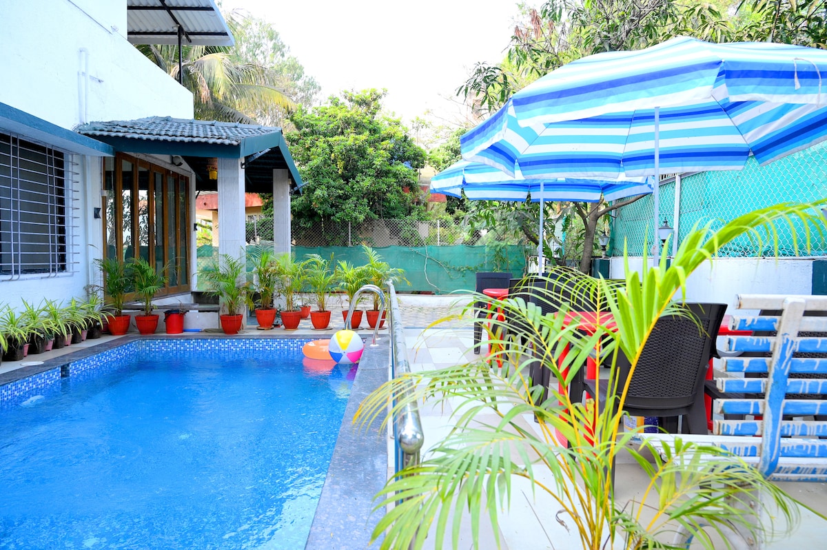 A private swimming pool is surrounded by vibrant green plants and colorful lounge chairs. Covered areas offer shade, while a few inflatable toys float in the water. The space is designed for relaxation in a tranquil environment.