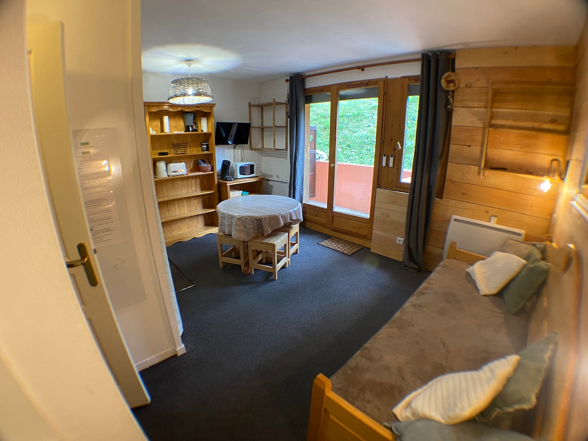A cozy living space features wood accents and a warm colour palette. A spacious bed is accompanied by a pull-out couch. A wooden table occupies the center, while a television and microwave are visible on a shelf. Large glass doors open to a balcony area.